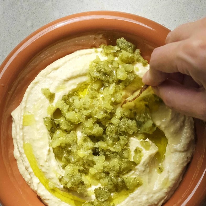 Krämig hummus toppad med rivna frysta cornichons och olivolja, serverad med chips.