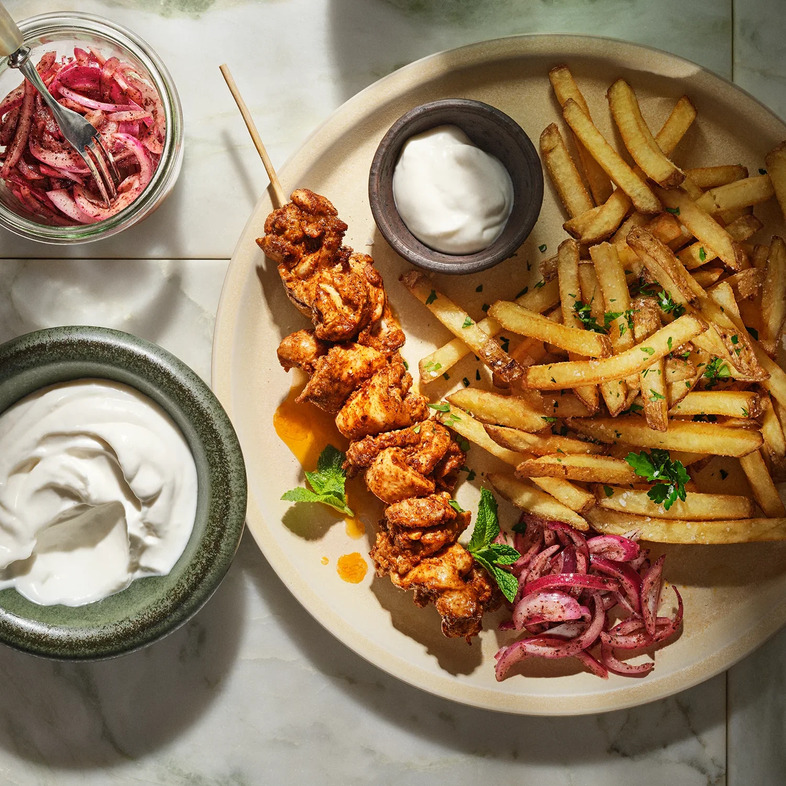 Grillade shawarmaspett med kyckling serverade med krämig toum, sumaclök och pommes frites på ett fat.