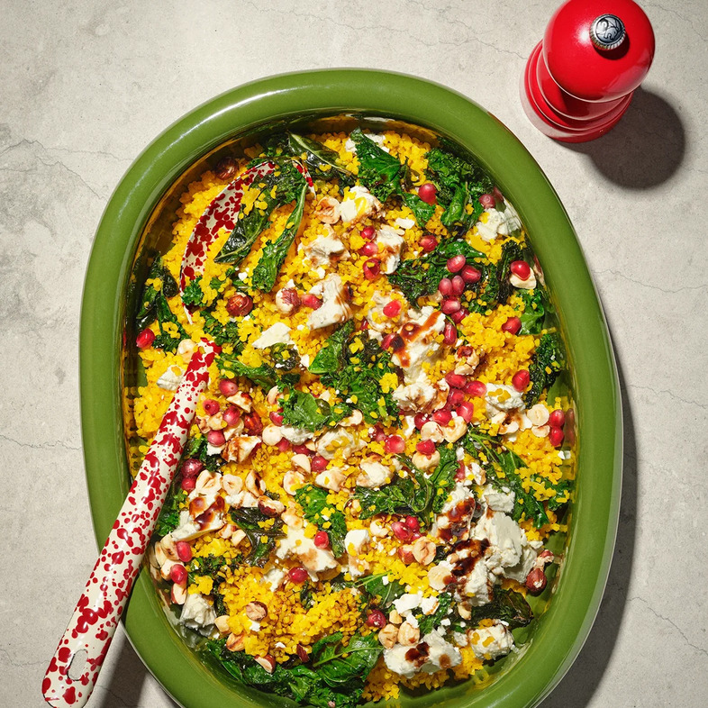 Ett stort serveringsfat med gyllengul bulgur blandad med grönkål, toppad med smulad fetaost, rostade hasselnötter och rubinröda granatäppelkärnor.